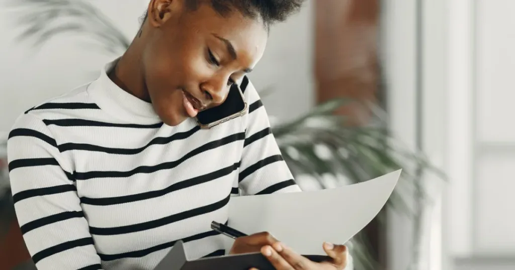 African woman on a phone call while taking notes, managing electricity, cable and internet bill payments