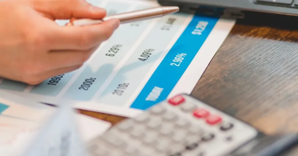 Close-up of a hand using a pen and calculator to compare interest rates on a chart