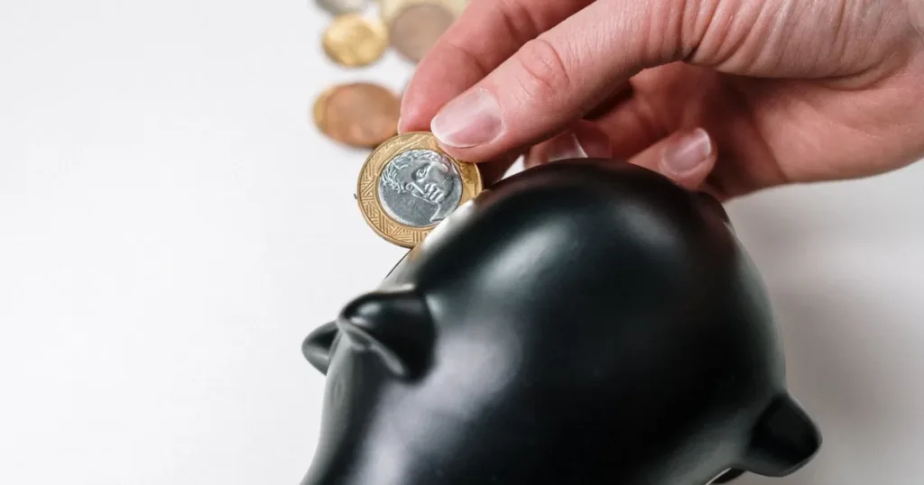 Hand placing a coin into a piggy bank, representing the choice between using savings or taking an emergency loan.