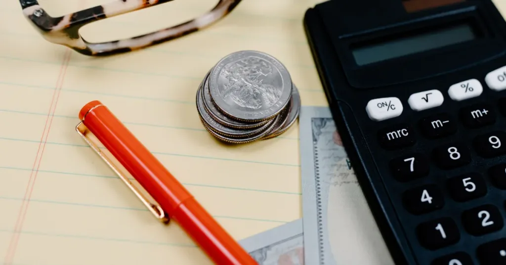 Notebook, red pen, calculator and coins laid out on a desk for building a monthly money plan