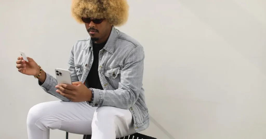 A young Nigerian man with an afro hairstyle holding a bank card while using his smartphone