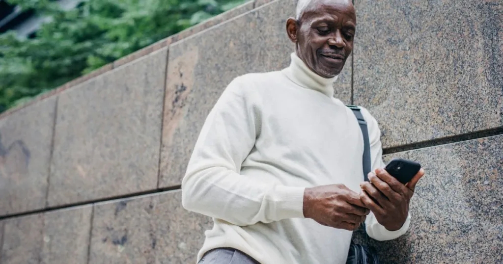 Cheerful young man browsing a mobile banking app on his smartphone on the street