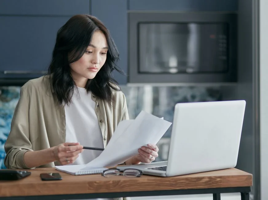 Woman reviewing financial documents and working on a laptop at a home office desk