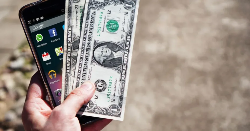 A hand holding US dollar banknotes next to a smartphone outdoors, representing money sent from abroad