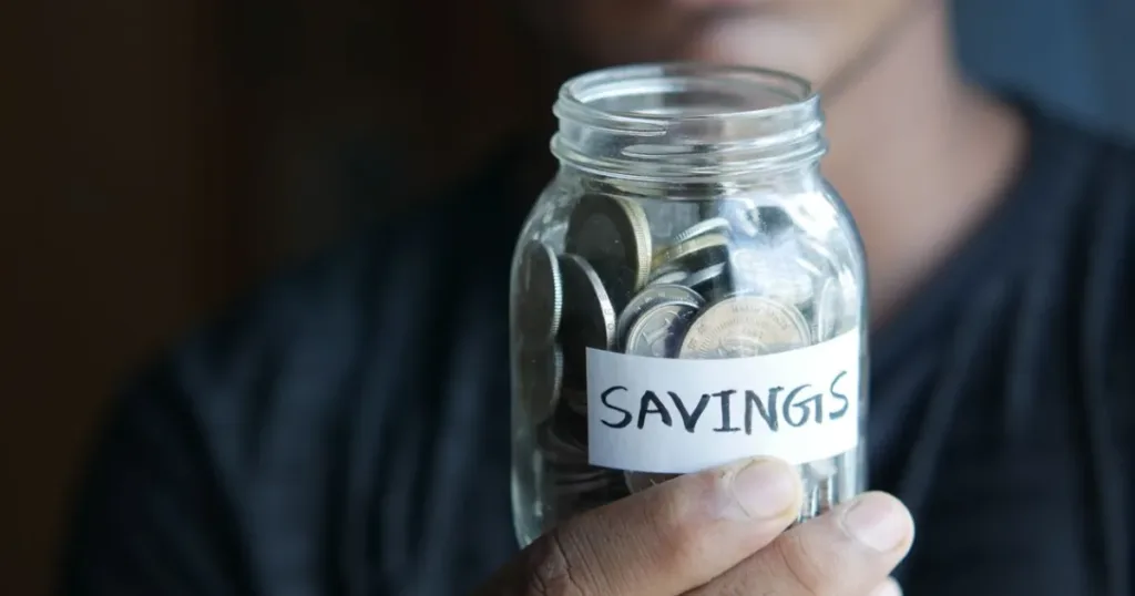 Hand holding a glass jar full of coins labeled Savings