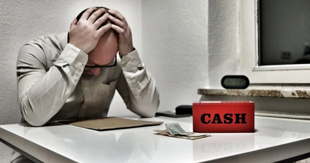 Stressed man holding his head at a desk with a cashbox and money