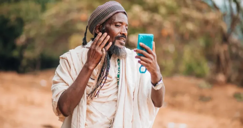 A man in traditional African attire using a smartphone outdoors, representing mobile payment apps