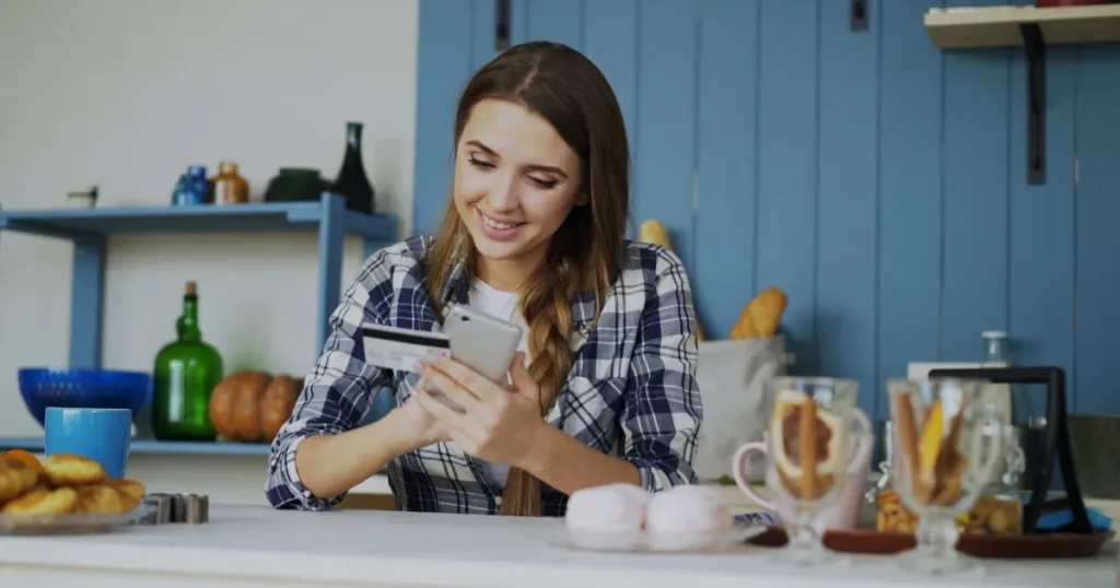 Smiling woman using a smartphone with a credit card in hand, cozy indoor kitchen setting